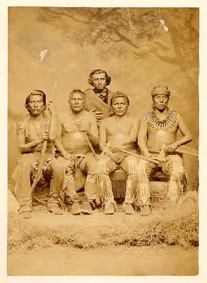 Orang Pawnee — Portrait of four seated Pawnee chiefs, all wearing decorated leggings, moccasins and peace medals, with a white man in the background dressed in a three-piece suit.
